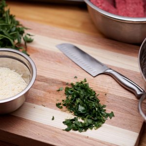 Auf einem Holzschneidbrett liegt ein Messer mit geschnittenen Kräutern und eine Metallschüssel mit Käse