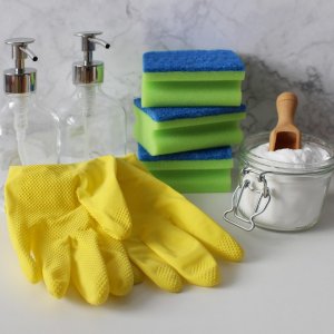 Auf einer Ablage liegen gelbe Gummihandschuhe, grüne Putzschwämme, zwei Pumpflaschen und ein Glas mit Sodapulver