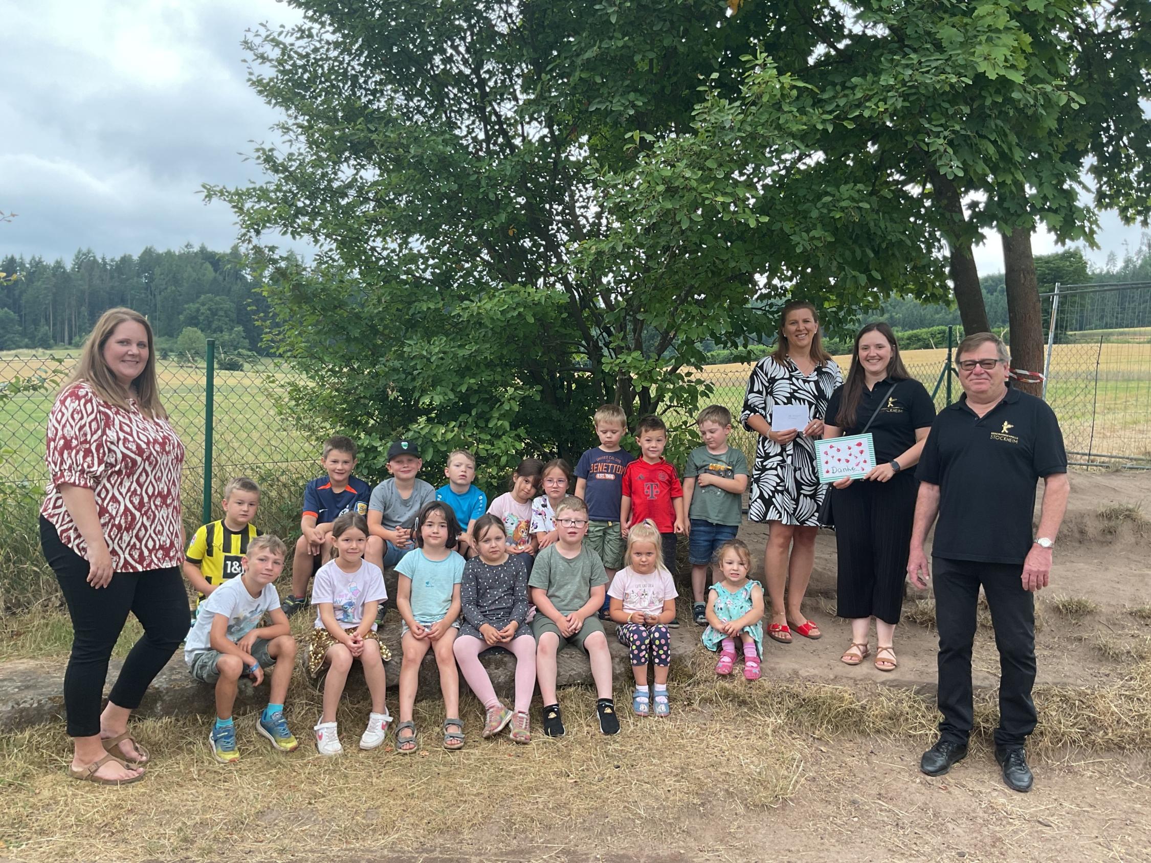 Einige Kinder sitzen auf Steinen und die Erzieher und die Vertreter der Kapelle stehen daneben
